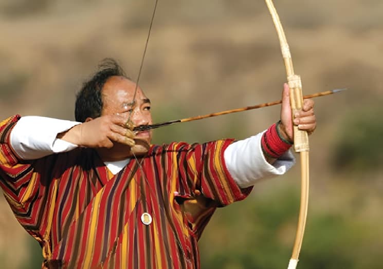 An archer in a striped gho drawing a traditional bamboo bow